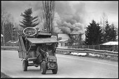 Tractor piled high with belongings on highway, smoke from burning buildings billowing up in background